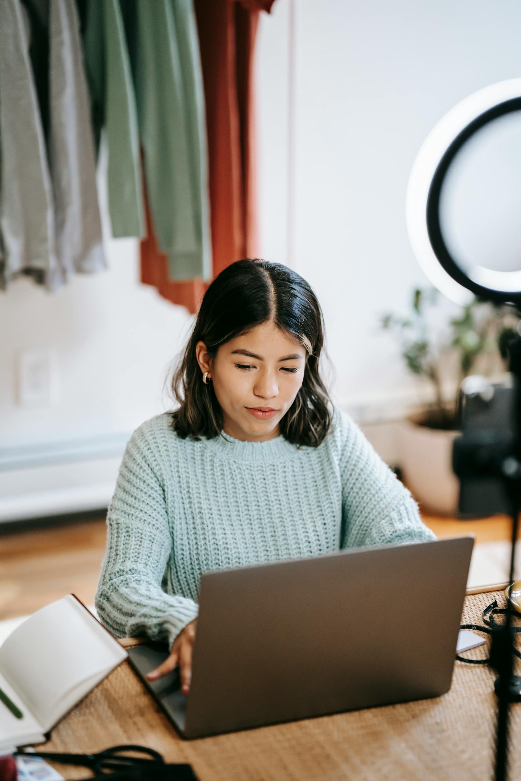 Focused young woman using laptop at home office with casual attire and bright lighting.